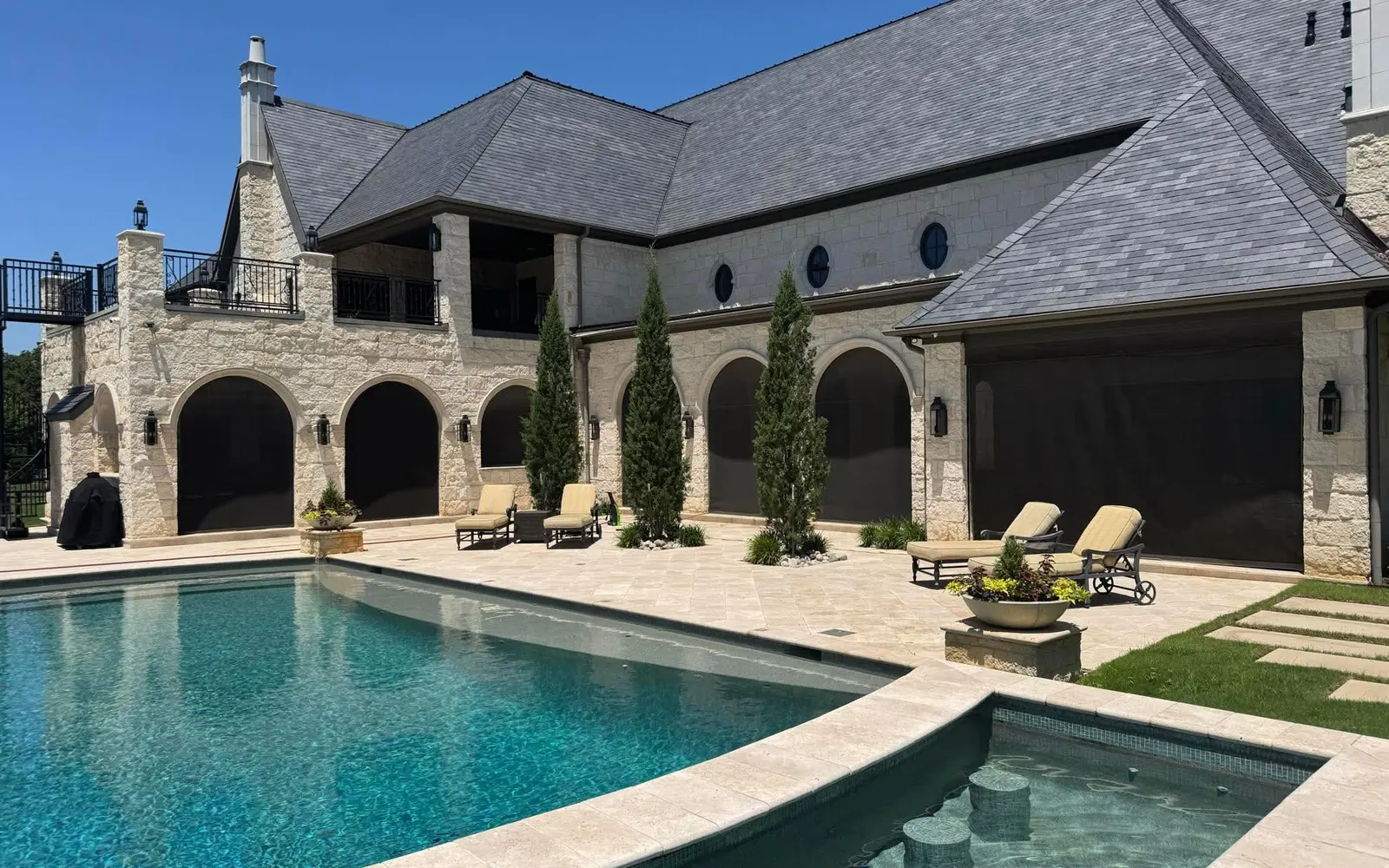 Large stone house and pool with dark motorized patio screens across arched openings, from EX Design Group